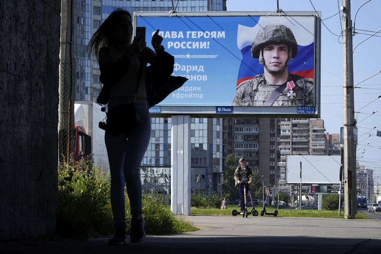 A woman is seen in silhouette looking at her cellphone with a poster glorifying a Russian soldier in the background.