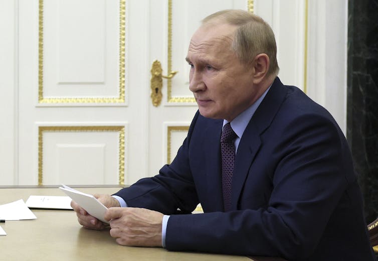 Vladimir Putin sits at a desk holding papers with gilded doors behind him.