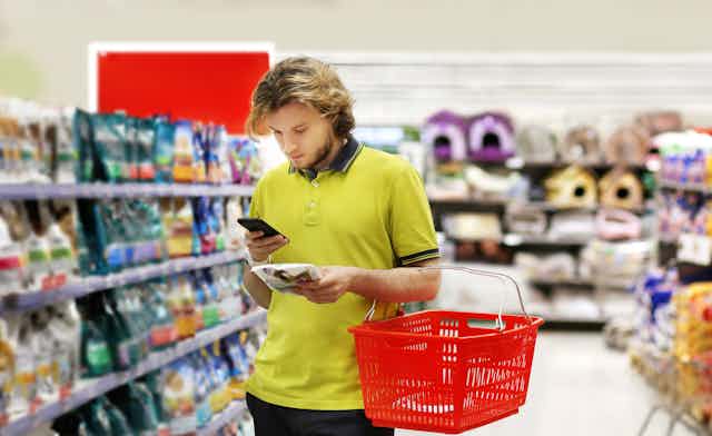 Man in yellow shirt in supermarket carrying red shopping basket looking at his phone