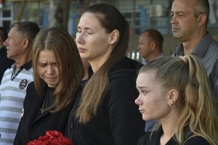 Three young women in black weep, one of them with her head bowed.