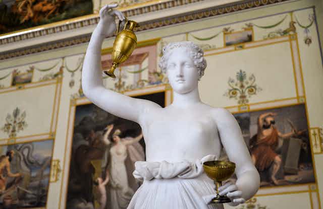 White marble sculpture of a bare-breasted woman lifting a gold urn and holding a cup, in the fashion of the goddess Hebe.