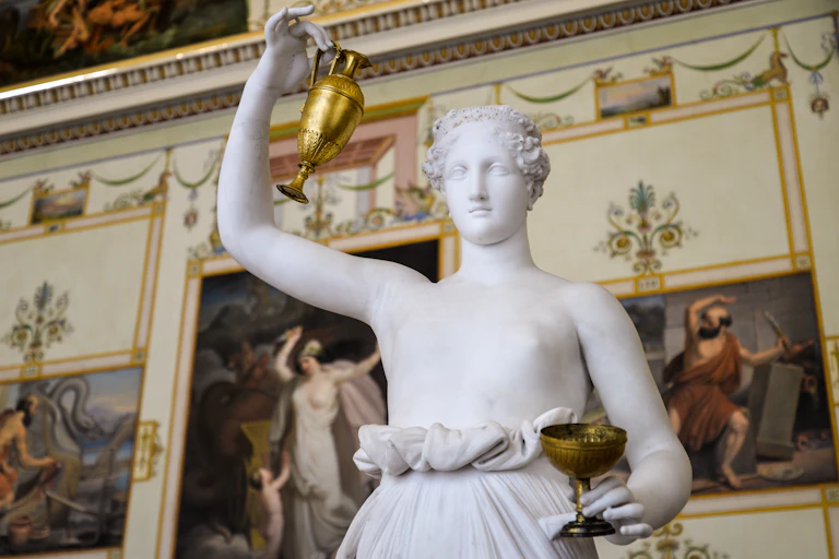 White marble sculpture of a bare-breasted woman lifting a gold urn and holding a cup, in the fashion of the goddess Hebe.