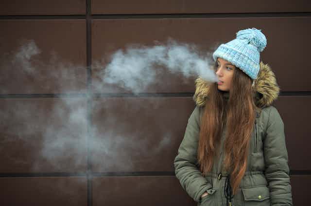 Wearing an overcoat and wool cap on a cold day, a teenage girl standing on the street smokes an electronic cigarette.
