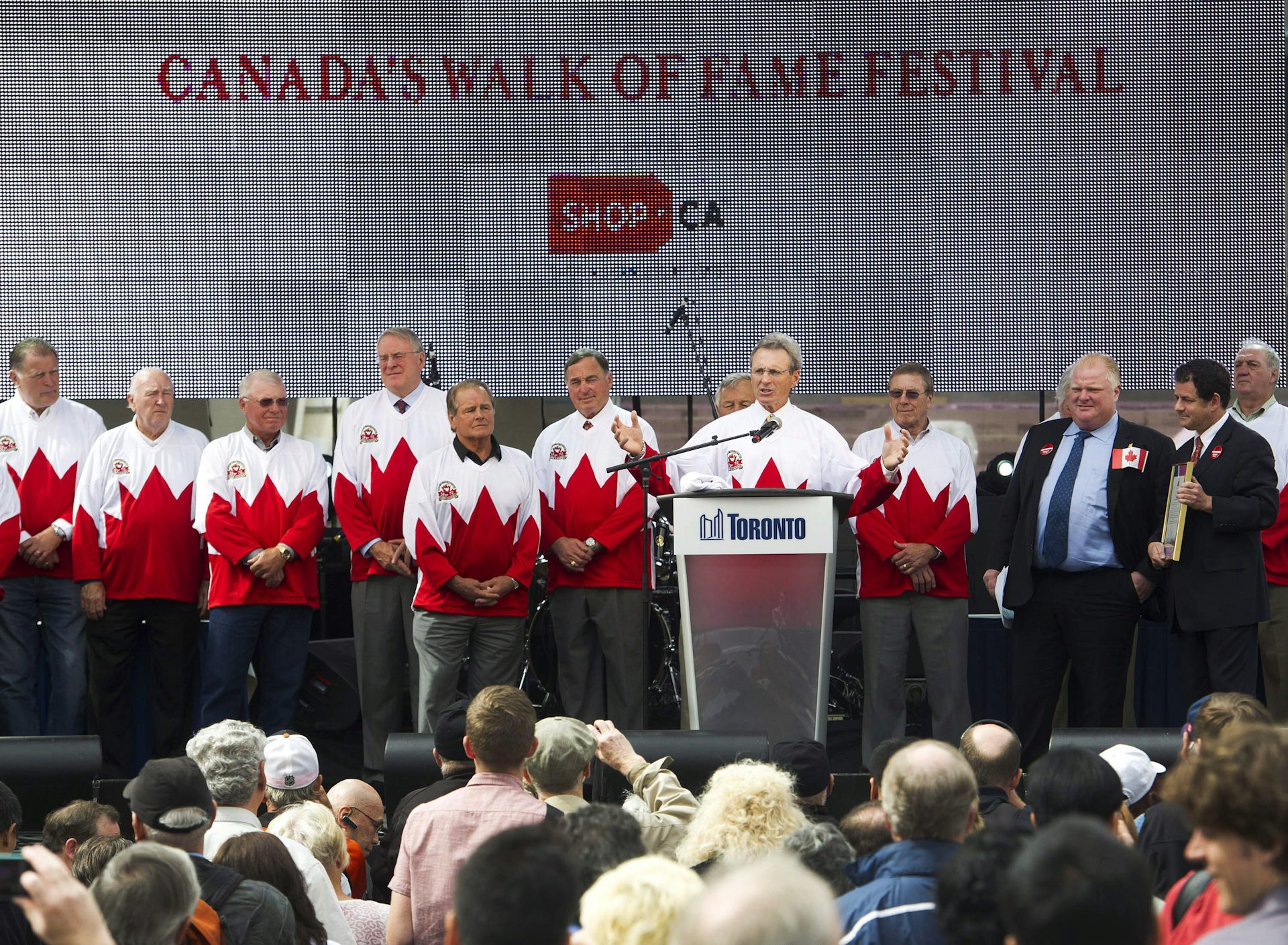 A man, standing on a stage with several other men in matching Canadian maple leaf sweaters, speaks from behind a podium.