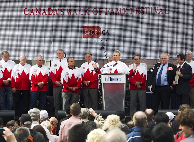 A man, standing on a stage with several other men in matching Canadian maple leaf sweaters, speaks from behind a podium