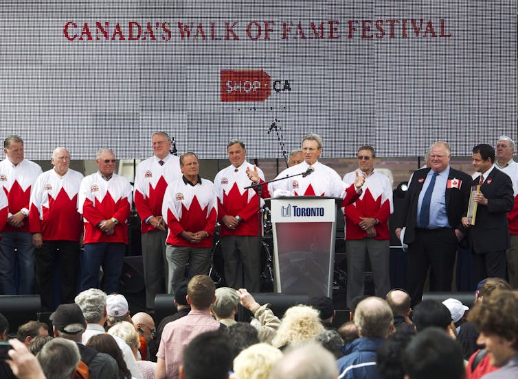A man standing on a stage with several other men in matching Canadian maple leaf sweaters speaks from behind a podium
