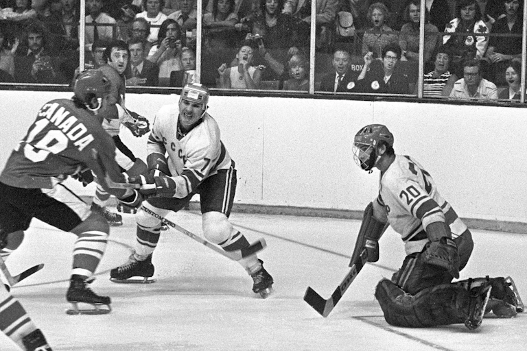 A black and white photograph of a Canadian hockey player shooting a puck at a U.S.S.R. goalie
