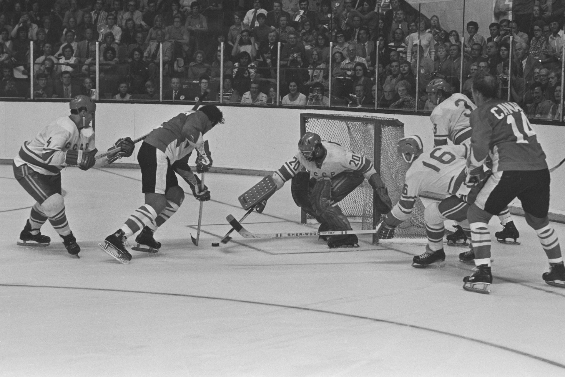 A black and white photo of Canadian hockey player about to score a goal on a U.S.S.R. goalie while his teammate tries to stop it with his hockey stick