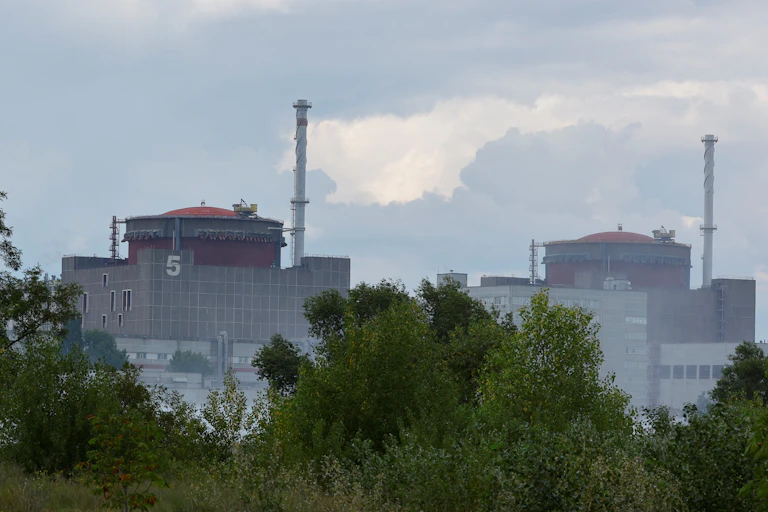 A row of small trees and shrubs in the foreground; in the background, three large cylindrical towers surrounded by massive concrete structures.