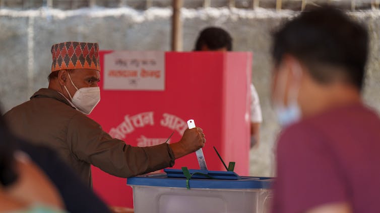 A man in a colourful fez and wearing a mask pushes a ruler inside a ballot box,