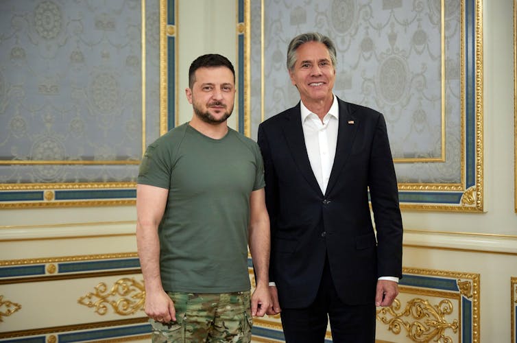 Zelensky in military fatigues and Blinken in a suit, smiling for cameras.