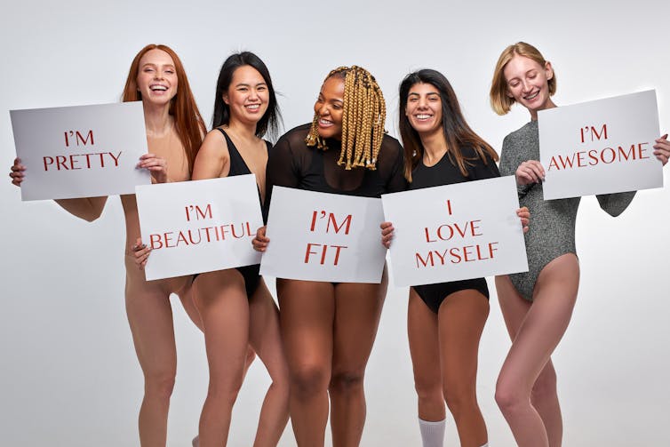 Sekelompok model perempuan yang mengenakan korset berpose dengan memegang tanda dengan pesan positif tentang tubuh
.