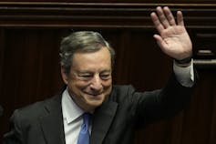 A man with grey-ish hair and wearing a suit smiles and waves.