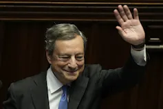 A man with grey-ish hair and wearing a suit smiles and waves.