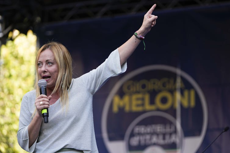 A blonde woman speaks into a microphone and points upwards. A sign that says Meloni is seen behind her.