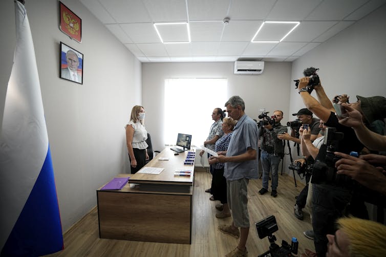 A group of people stand together in a room facing a portrait of Vladimir Putin, reading from a paper while journalists take photos and videos behind them.