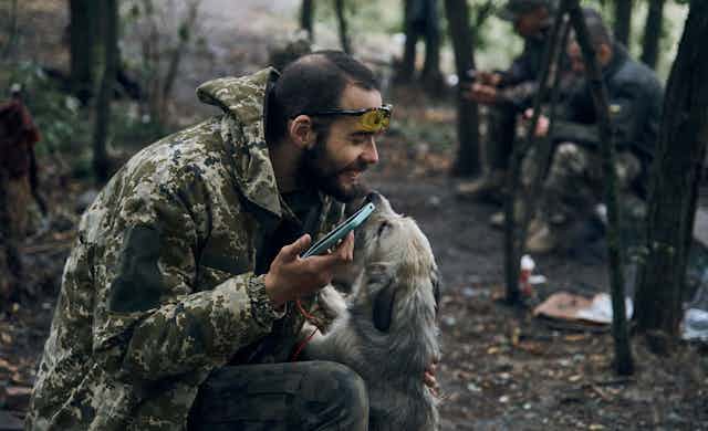 A soldier holding a smartphone smiles as he pats a shaggy dog that licks his face.