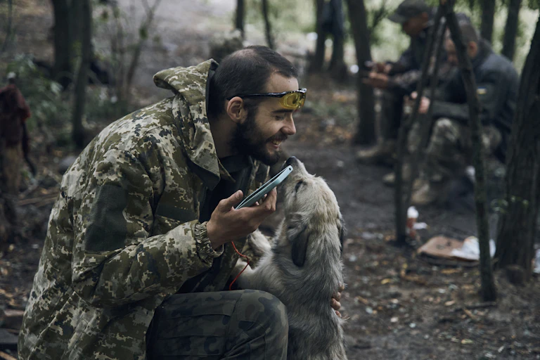 A soldier holding a smartphone smiles as he pats a shaggy dog that licks his face.