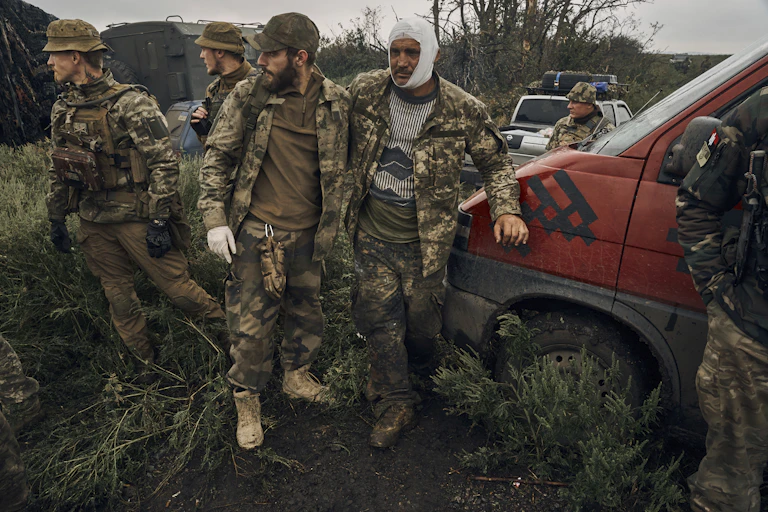Camouflage-clad men, one with bandages around his head, stand in brush near vehicles.