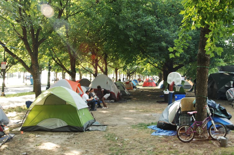 Tents line a leafy park; some people can be seen chatting outside one tent