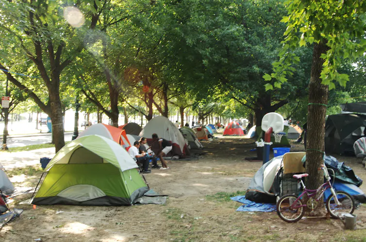 Tents line a leafy park; some people can be seen chatting outside one tent