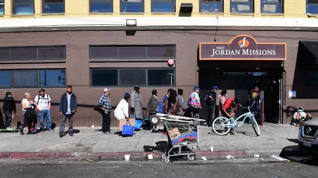 People of all races line up on a sidewalk outside the Jordan Missions; a shopping cart full of personal belongings is in the foreground