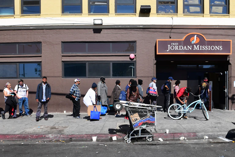 People of all races line up on a sidewalk outside the Jordan Missions; a shopping cart full of personal belongings is in the foreground