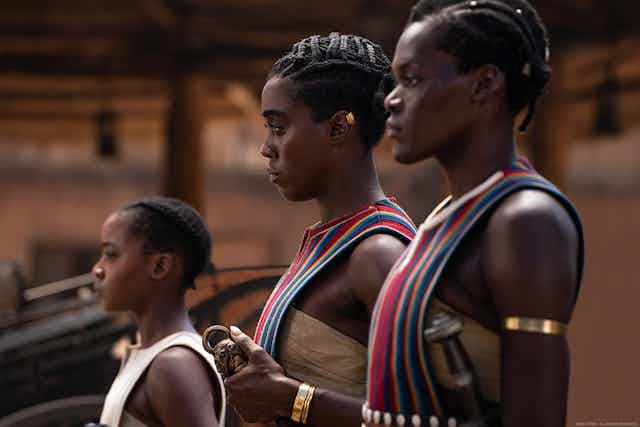 Two women and a girl stand in profile at attention. They are dressed in beaded attire with traditional African braided hairstyles