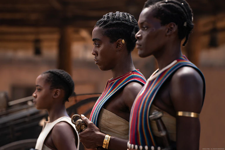 Two women and a girl stand in profile at attention. They are dressed in beaded attire with traditional African braided hairstyles