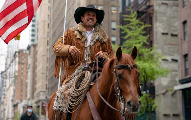 A white man wearing a black cowboy hat and a brown jacket rides a brown horse and holds an American flag in an empty Manhattan street