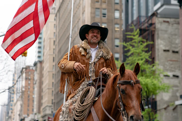 A white man wearing a black cowboy hat and a brown jacket rides a brown horse and holds an American flag in an empty Manhattan street