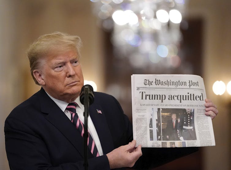 A white older man with yellow hair and a suit holds up a Washington Post newspaper that says 'Trump acquitted' in large black font