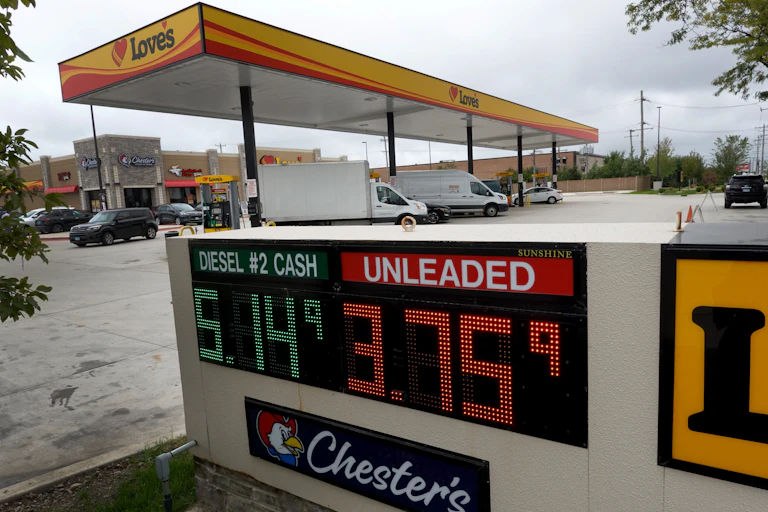 A gas station is shown with cars and vans in the background and a display in the front showing prices.