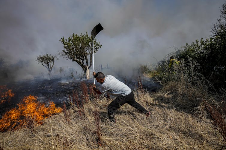 A man beats at flaming grasses with a shovel. Fire-blackened landscape is in the background.