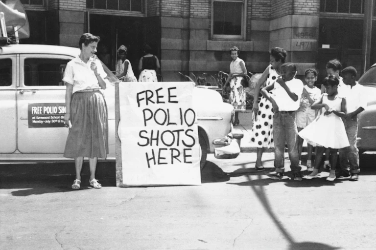 An historical photo of a group of children, outside a public school, standing by a sign that says "Free Polio Shots Here."