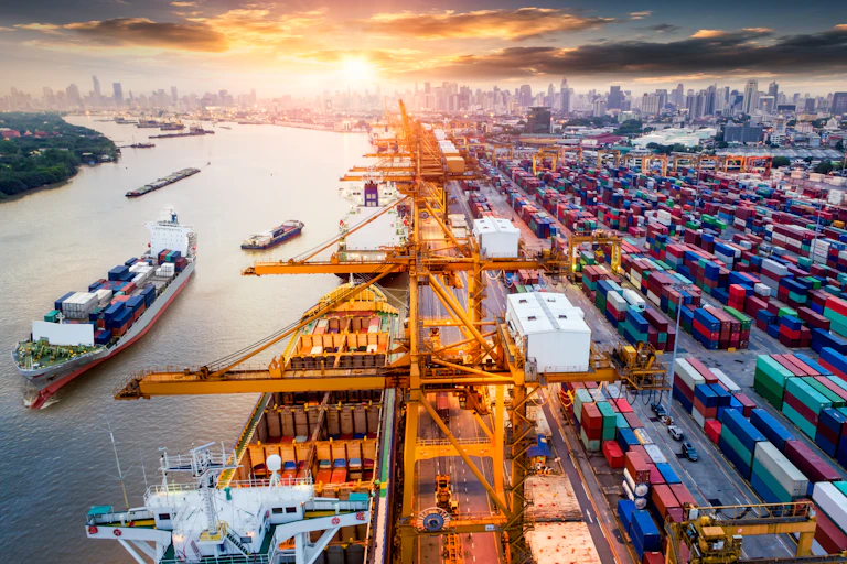 A busy port in Thailand with a city in the background, ships on the water and lots of shipping containers in port.