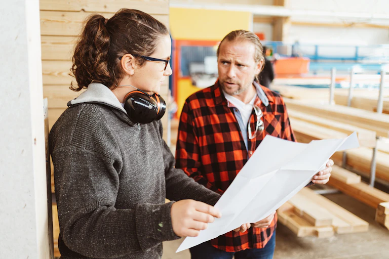 Two people talk to one another at a construction site while looking at a blueprint