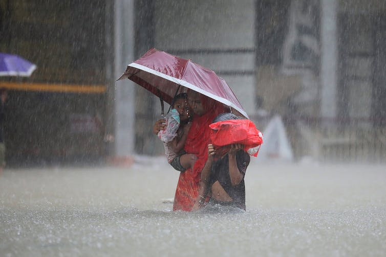 Two children cling to a woman as they walk through nearly waist-deep water on a city street in pouring rain.