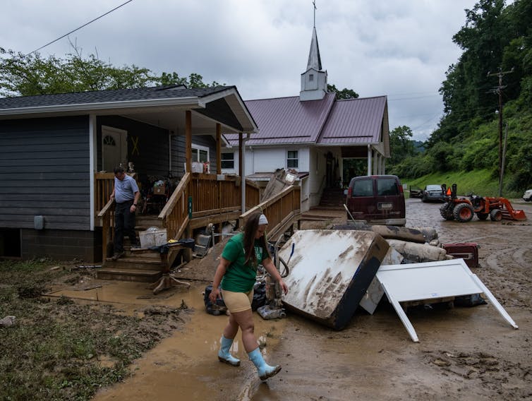A girl in rain boots walks through a mud-filled yard. Damaged mattresses and other belongings from a flooded house are piled nearby.