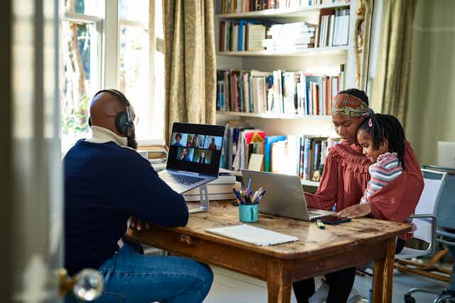 a man wearing headphones looks at a video conference call screen on a laptop while across the table a woman with a young child in her lap looks at a laptop screen