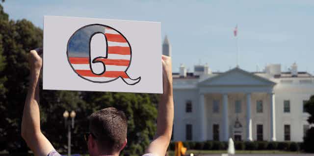 A man stands in front of the White House holding a sign with the letter Q in the colours of the US flag.