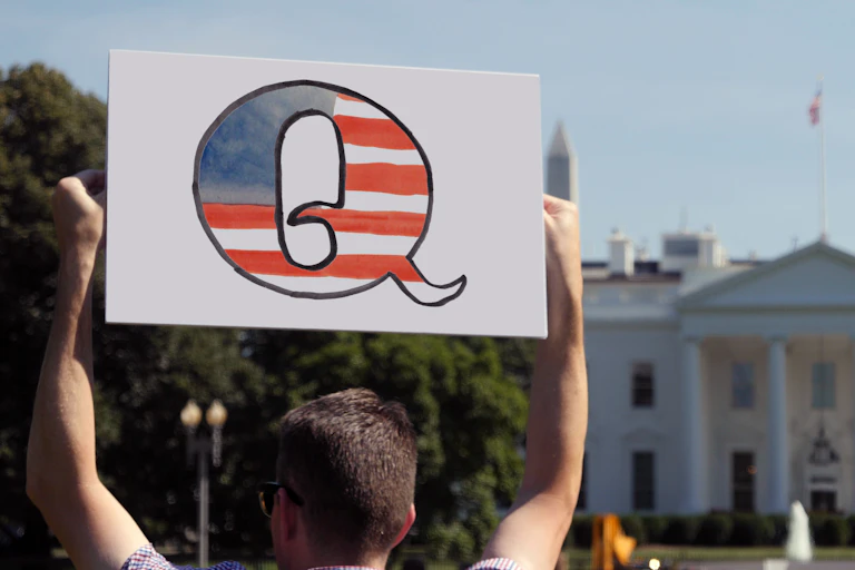 A man stands in front of the White House holding a sign with the letter Q in the colours of the US flag.