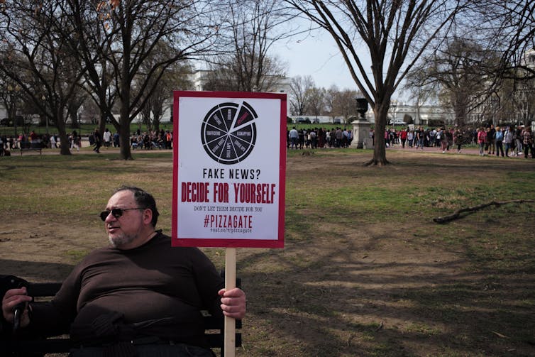 A man sits on a bench in a park holding a sign that reads: fake news decide for yourself Pizzagate.