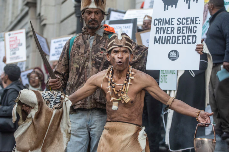 A protest outside a grand old building, one placard reading 'Crime scene: river being murdered". In front, a man in traditional Khoi attire.