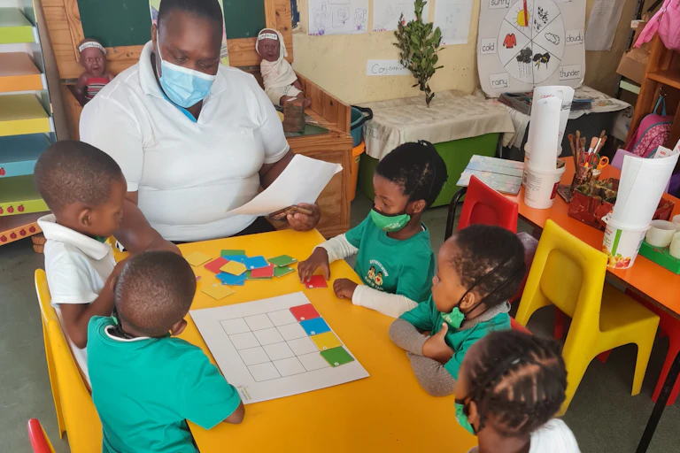 A group of five young children, some of them wearing face masks, sit around a yellow table with a woman who is pointing to colourful puzzle pieces.