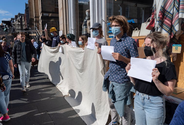 A line of people in face masks holding up blank pieces of paper, several are also holding a large, blank banner.