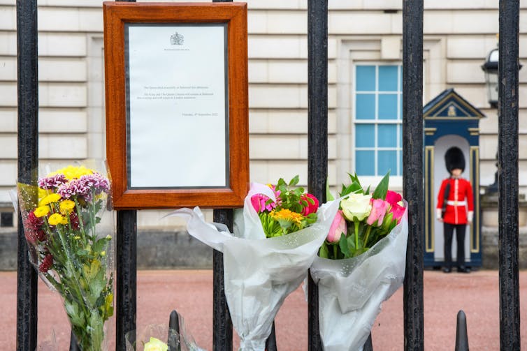 A white poster in a wooden frame attached to metal gates with bunches of flowers.