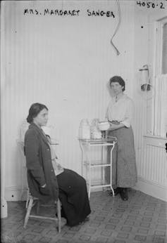 A woman sits in a chair being tended to by a nurse, standing