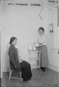 A woman sits in a chair being tended to by a nurse, standing