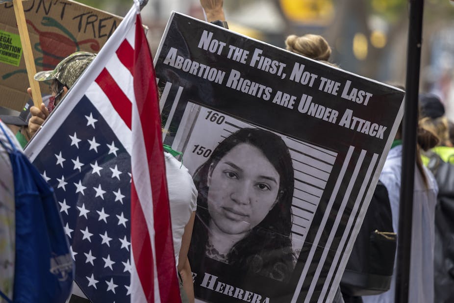 Protest sign saying 'Not the first, not the last abortion rights are under attack' with the mugshot of Lizzie Herrera; an American flag is in the foreground